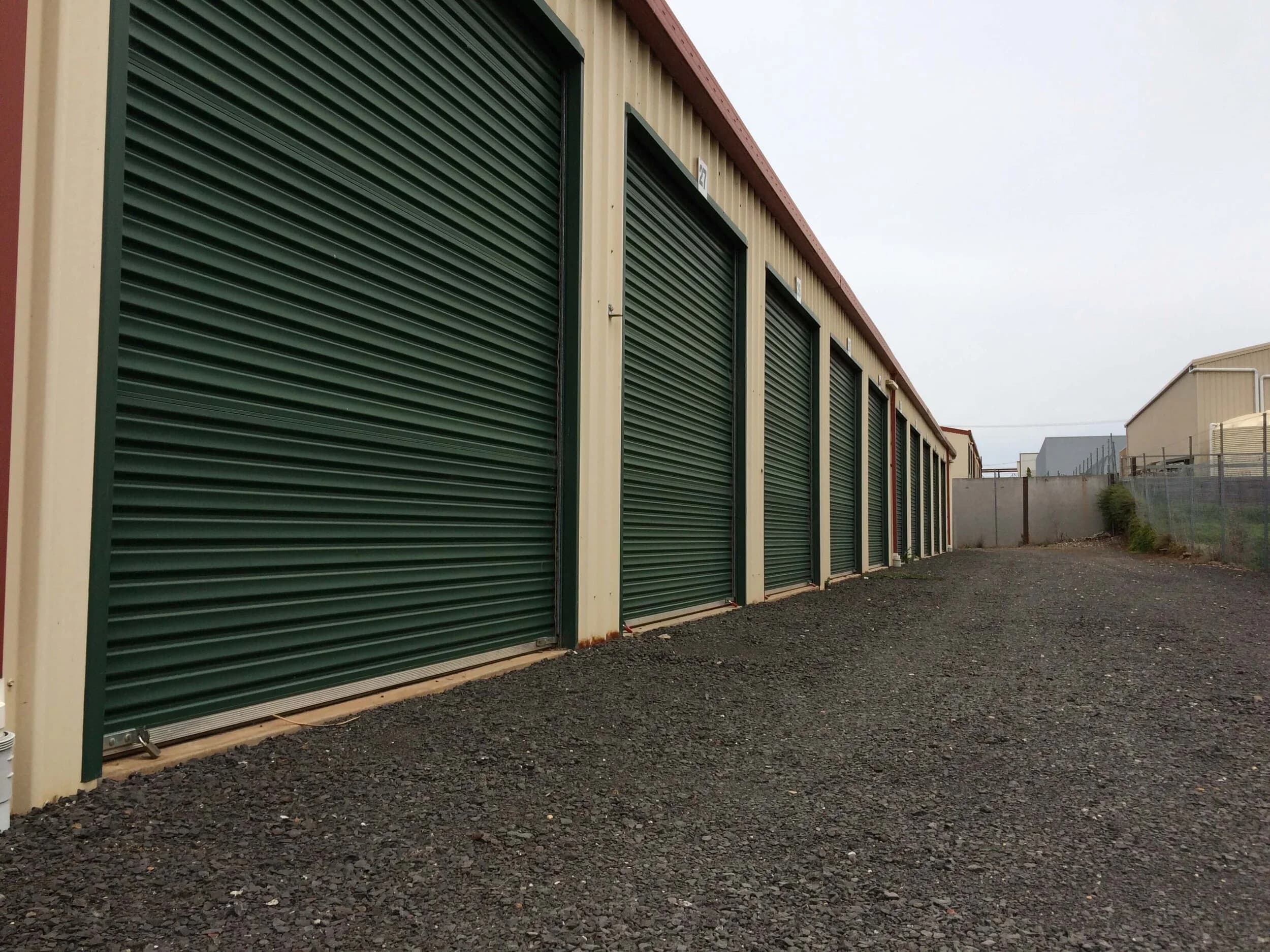 A long row of green roller-door units with a wide gravel driveway
